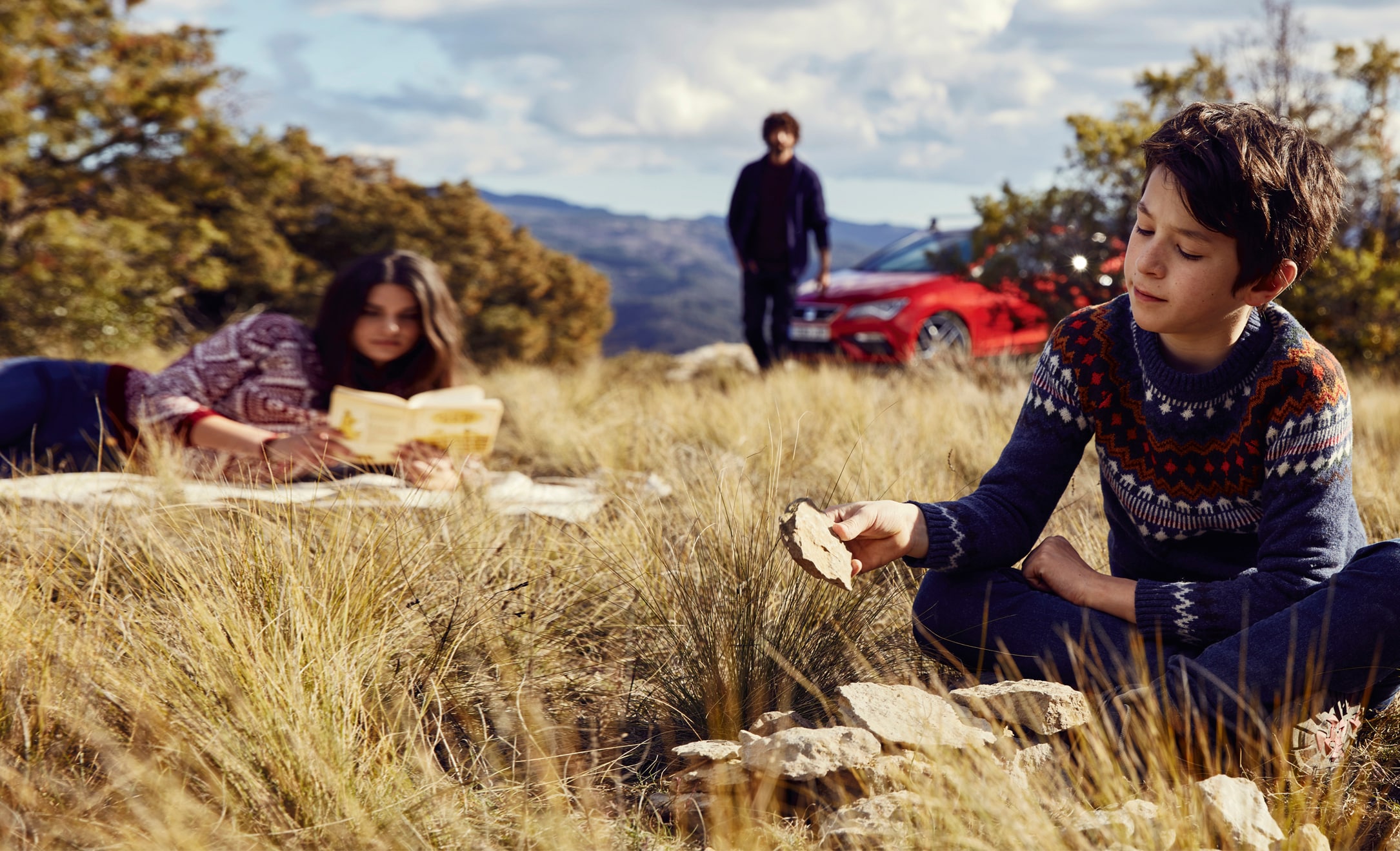 A family of three relaxing in a field, with their red SEAT Leon parked in the background.