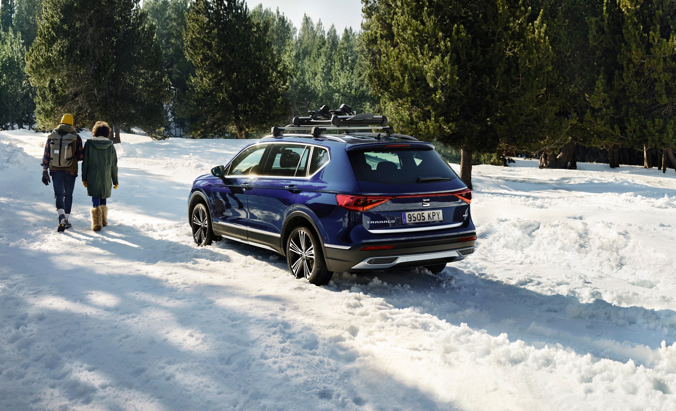 Side rear view of a blue SEAT Tarraco with roof bars in the snow, two people walking beside it.