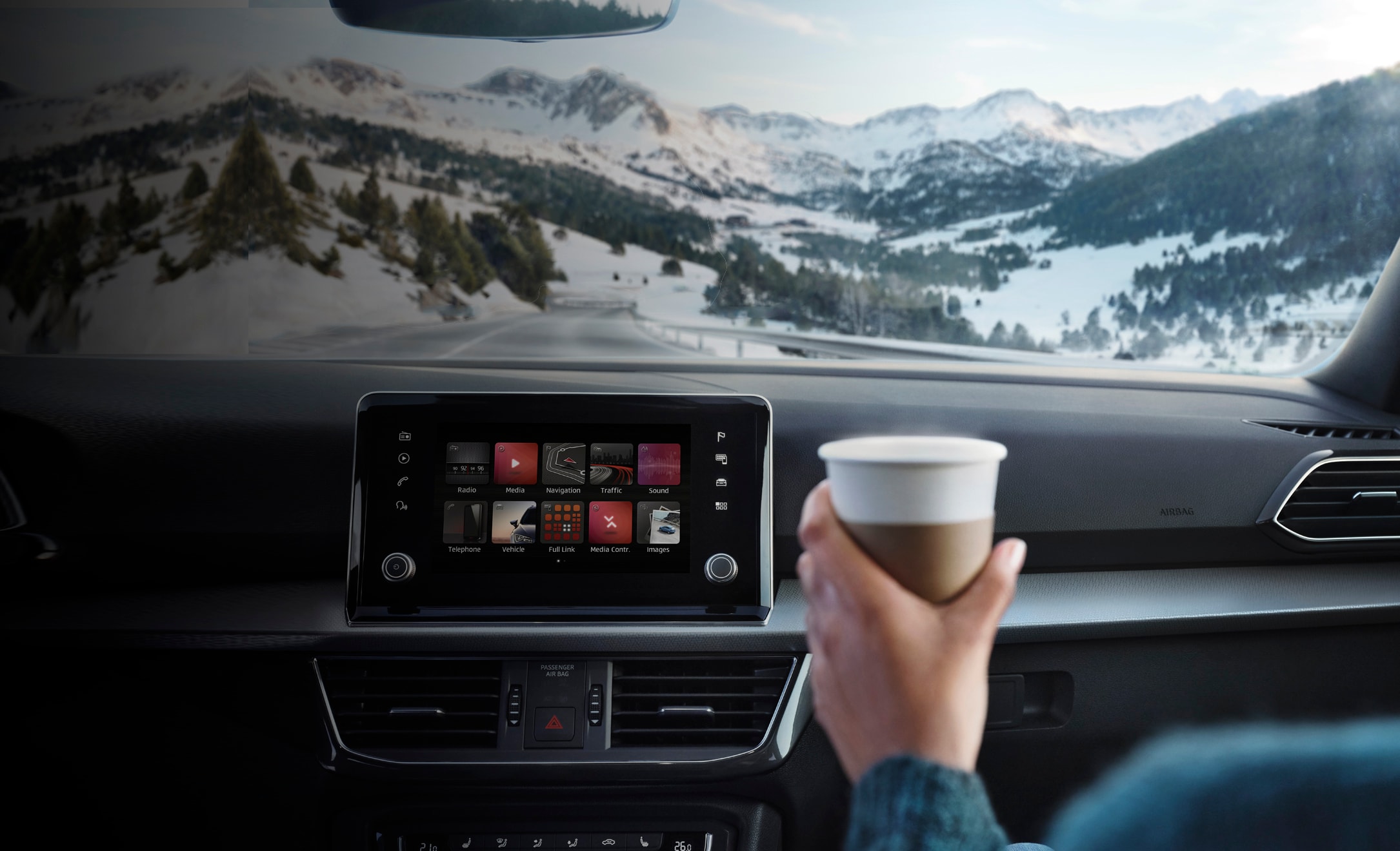 A man holding a coffee cup in front of a SEAT car’s infotainment system, with a view of snowy mountains through the front windscreen.