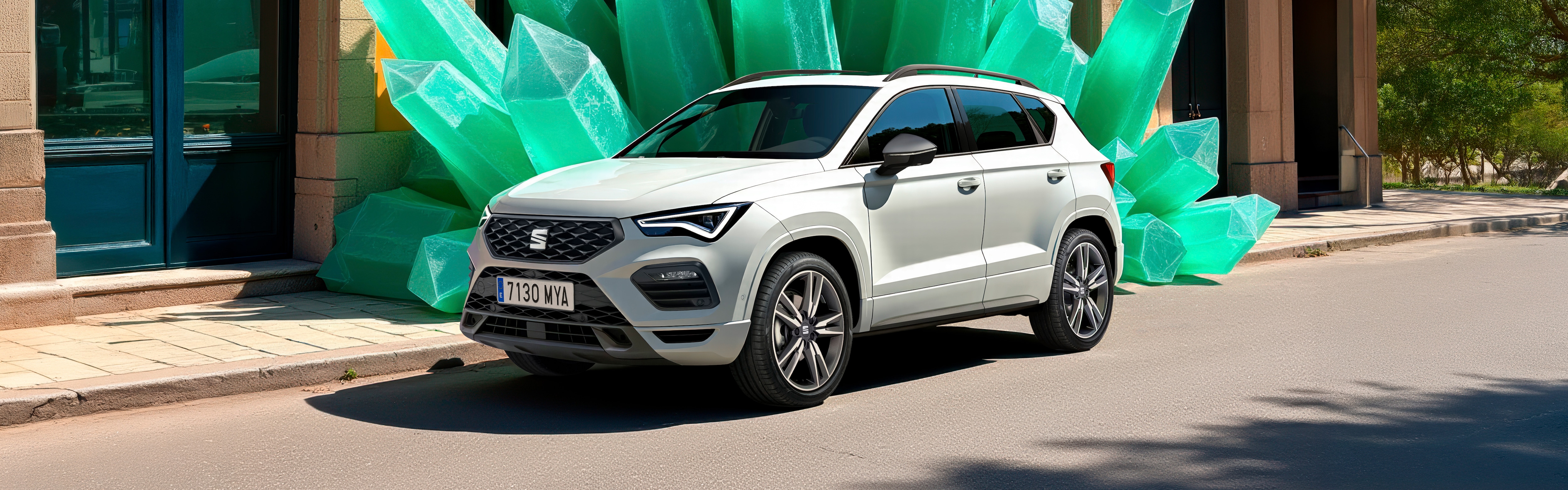 A white SEAT Ateca SUV parked on a street in front of an orange building with arched windows, with a large green stone structure resembling giant crystals behind it. The scene is bright and sunny with clear blue skies.