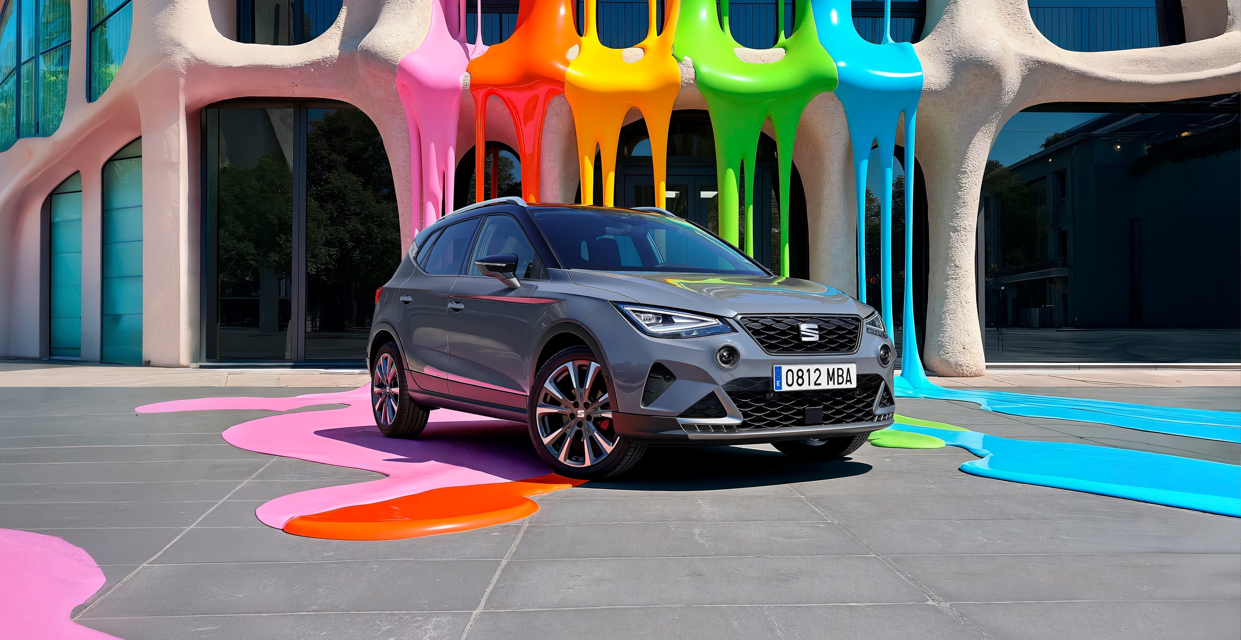 A SEAT Arona compact SUV parked in front of a colourful, artistic building with large irregular windows and vibrant paint drips in pink, orange, yellow, green, and blue flowing down the façade and onto the ground. The car's license plate reads '0812 MBA