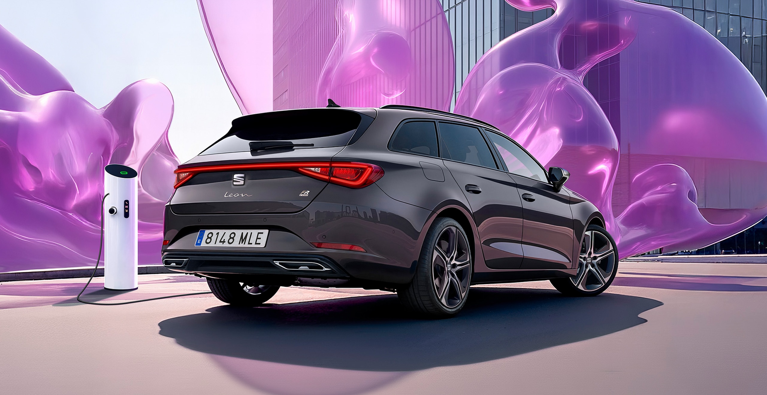A SEAT Leon Sportstourer parked near an electric vehicle charging station in an urban setting, with a tall glass building in the background and large abstract purple sculptures surrounding the area. 