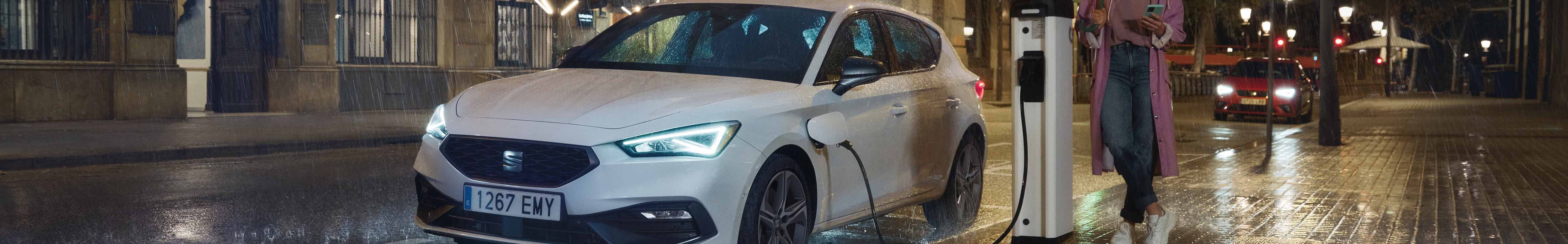 Hybrid car seat leon charging at a public charging station on a a rainy street at night, highlighting the advantages of hybrid vehicles.